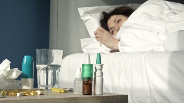Grip, soğuk algınlığı ya da koronavirüs belirtileri gösteren hasta bir kadın yatakta yatıyor. Ön planda bir komodin var. Üzerinde bir sürü farklı ilaç var.. — Stok video