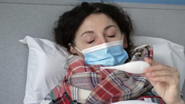 Donna malata di mezza età con una maschera protettiva si trova a letto e misura la temperatura. Ha sintomi di influenza, raffreddore o coronavirus. — Video Stock