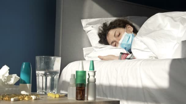 Zieke vrouw met een medisch masker op het gezicht met symptomen van griep, of coronavirus ligt in bed. Op de voorgrond staat een nachtkastje waarop veel verschillende medicijnen staan. — Stockvideo