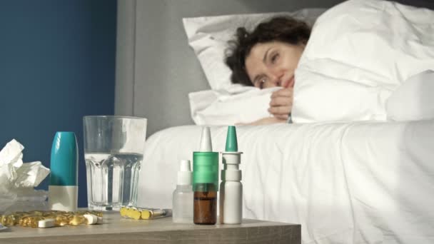 Beteg nő tünetei influenza, hideg vagy coronavirus fekszik az ágyban. Az előtérben van egy éjjeliszekrény, amelyen sok különböző gyógyszer van.. — Stock videók