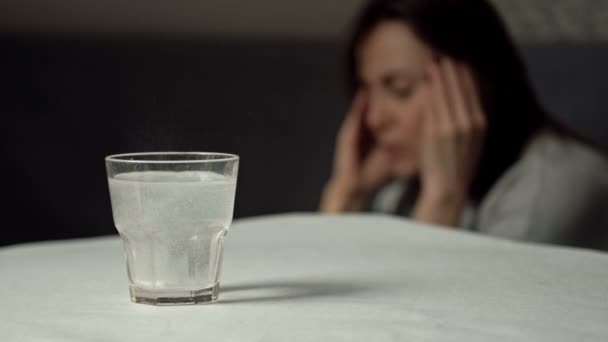 Verre d'eau avec un comprimé dissolvant. Gros plan. En arrière-plan, une femme pénible se frotte les tempes. — Video