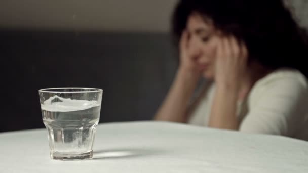Ett glas vatten med en upplöst tablett. Närbild. I bakgrunden gnuggar en plågad kvinna sina tempel.. — Stockvideo