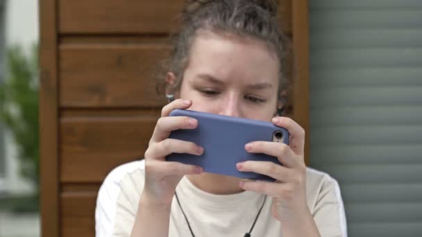 Έφηβη κοπέλα που κρατάει το τηλέφωνο παθιασμένη με τις εφαρμογές κοινωνικής δικτύωσης smartphone κάθεται μόνη της στο σπίτι. Τεχνολογία εθισμένος έφηβος παίζει κινητά παιχνίδια, γραπτών μηνυμάτων, βλέποντας βίντεο σχετικά με τη σύγχρονη τεχνολογία gadget. — Αρχείο Βίντεο
