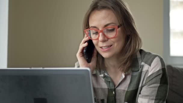 Femme d'affaires de bonne humeur. Belle femme d'âge moyen est assis devant un ordinateur portable et avoir du plaisir à parler à quelqu'un sur son téléphone mobile. — Video