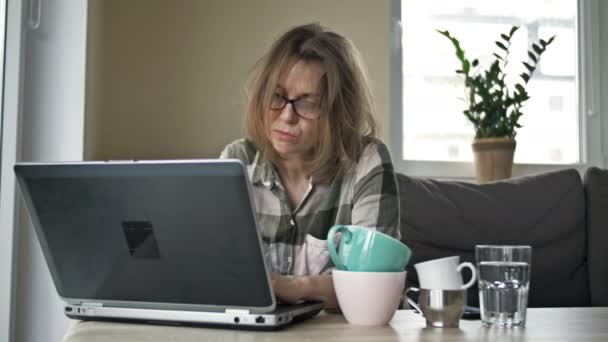 Mycket trött affärskvinna arbetar med bärbar dator hemma. Det finns flera tomma koppar te och kaffe på bordet. Kvinnan gnider sina trötta ögon och masserar sina tempel. Överarbetskoncept. — Stockvideo