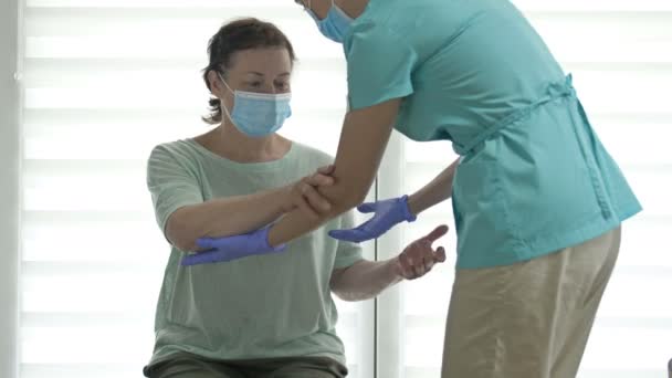 El médico o la enfermera están ayudando a una mujer mayor a ponerse de pie. Recuperarse de una lesión, cirugía o accidente cerebrovascular. Escudos faciales. Tratamiento durante la pandemia de Covid-19. — Vídeo de stock