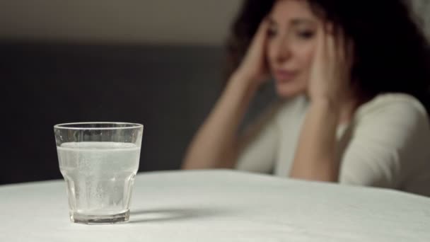 Un vaso de agua con una tableta disolvente. Primer plano. Al fondo, una mujer dolorida se frota las sienes. — Vídeos de Stock