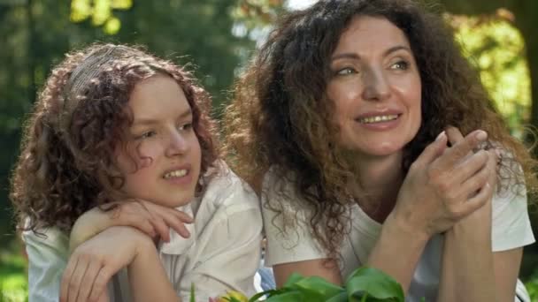 Retrato primaveral de mamá y su hija adolescente tumbada en la hierba. Ambos miran a la distancia y sonríen. Día de las Madres. — Vídeo de stock