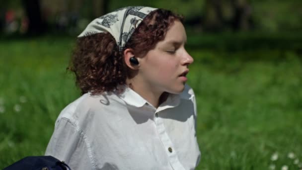Портрет красивої щасливої дівчини-підлітка, що сидить на траві, насолоджується сонячним днем, слухає музику з навушниками та співає . — стокове відео