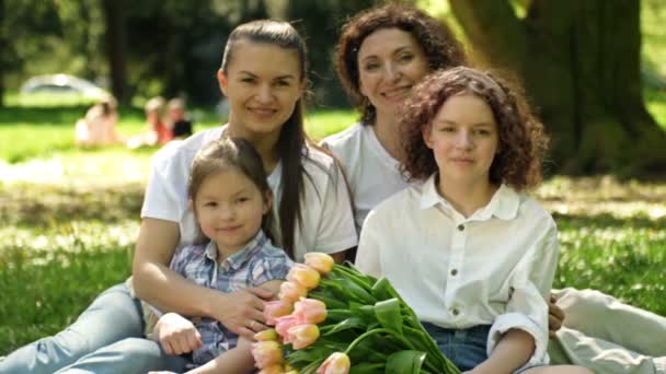 Dos mujeres con sus hijas están sentadas en el parque en la hierba. En primer plano hay un hermoso ramo de tulipanes. En el fondo de un parque de primavera. Día de las Madres. — Vídeo de stock