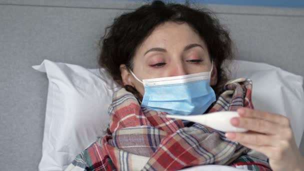 Koruyucu maskeli hasta orta yaşlı kadın yatakta yatıyor ve sıcaklığı ölçüyor. Grip, soğuk algınlığı ya da koronavirüs belirtileri var. Kadın sıcaklık ölçüm sonuçlarından rahatsız.. — Stok video