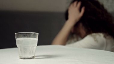 Çözünme tabletli bir bardak su. Yakın plan. Arka planda, acıyla boğuşan bir kadın şakaklarını ovuyor..