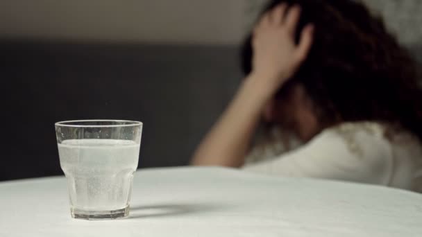 Un vaso de agua con una tableta disolvente. Primer plano. Al fondo, una mujer dolorida se frota las sienes. — Vídeo de stock