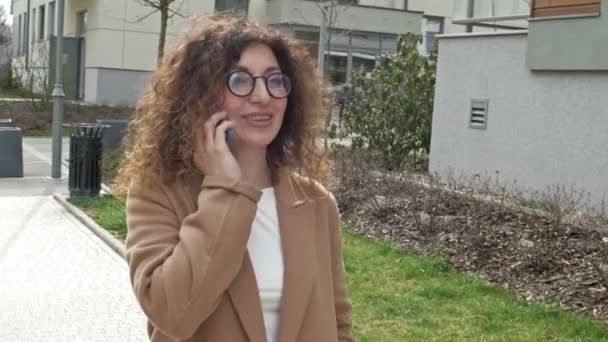 Een krullende vrouw van middelbare leeftijd met een bril loopt door een verlaten straat. Ze heeft plezier met iemand aan de telefoon.. — Stockvideo