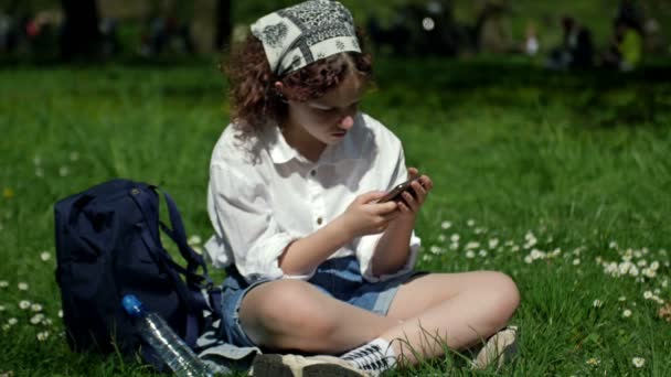 Schattig tienermeisje zit op het groene gras in het park en kijkt met belangstelling naar het scherm van haar smartphone. Naast haar staat een rugzak en een fles water.. — Stockvideo