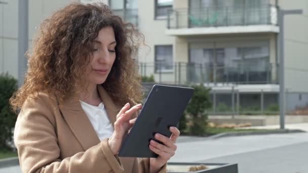 Mujer de pelo rizado de mediana edad pasea por una calle desierta de la ciudad. Se comunica con alguien a través de una tableta. — Vídeos de Stock
