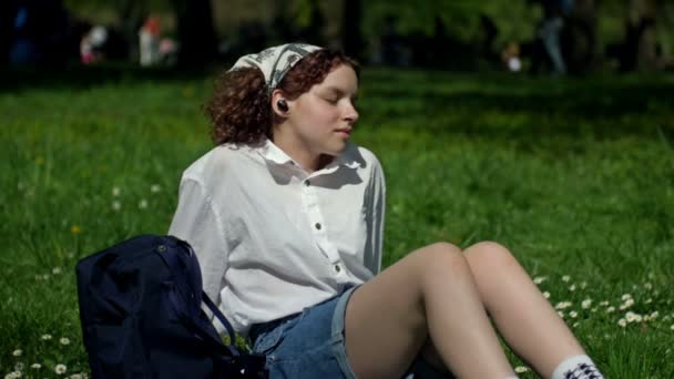 Chica adolescente se sienta en la hierba verde en el parque y escucha música, ojos cerrados, a través de auriculares. — Vídeos de Stock
