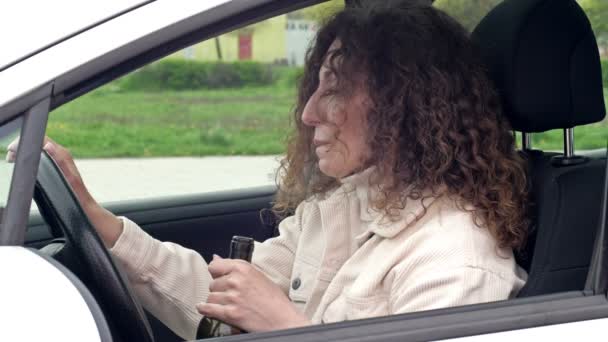 Bela mulher de meia-idade bebe vinho de uma garrafa sentada em seu carro. Ela já está muito bêbada.. — Vídeo de Stock