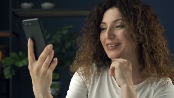 Belle femme de 40 ans avec un téléphone portable à la main sourit joyeusement lors d'un appel vidéo. On dirait qu'elle flirte avec quelqu'un.. — Video