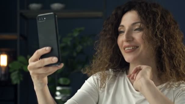 Belle femme de 40 ans avec un téléphone portable dans la main ondule et sourit joyeusement lors d'un appel vidéo. On dirait qu'elle flirte avec quelqu'un.. — Video