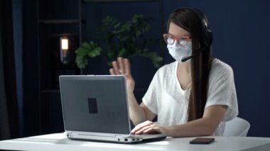 Coronavirüs salgını sırasında ofis işleri. Tıbbi maskeli bir iş kadını dizüstü bilgisayarın web kamerasına bakıyor ve biriyle konuşuyor..