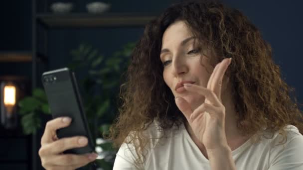Piękna 40-letnia kobieta z telefonem komórkowym w ręku uśmiecha się szczęśliwie podczas rozmowy wideo. Wygląda, jakby z kimś flirtowała.. — Wideo stockowe