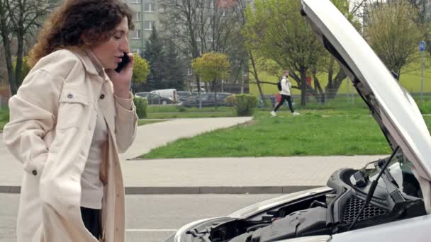 Orta yaşlı kadın sürücü arabasının açık kaputunun yanında duruyor. Telefonla birini arıyormuş. Yardıma ihtiyacı var gibi görünüyor.. — Stok video