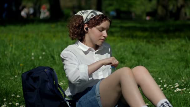 Милая школьница села отдохнуть на траве в весеннем парке. Девушка-подросток пьет воду из пластиковой бутылки и слушает музыку через беспроводной наушник. — стоковое видео