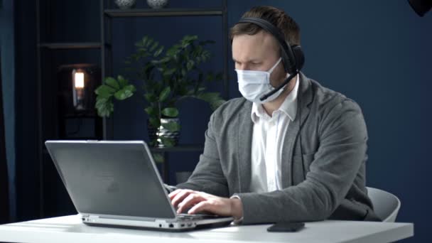 Kancelářská práce během epidemie koronaviru. Podnikatel středního věku ve sluchátkách pracuje s laptopem. Muž má na tváři ochrannou masku.. — Stock video
