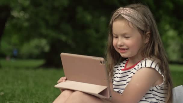 Funny girl 6-7 years old sits on the grass in a summer park with a tablet. Children learn gadgets easily. Modern technologies for children. — Stock Video