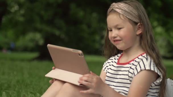 Vicces lány 6-7 éves ül a füvön egy nyári parkban egy tablettát. A gyerekek könnyen megtanulják a kütyüket. Modern technológiák gyermekeknek. — Stock videók