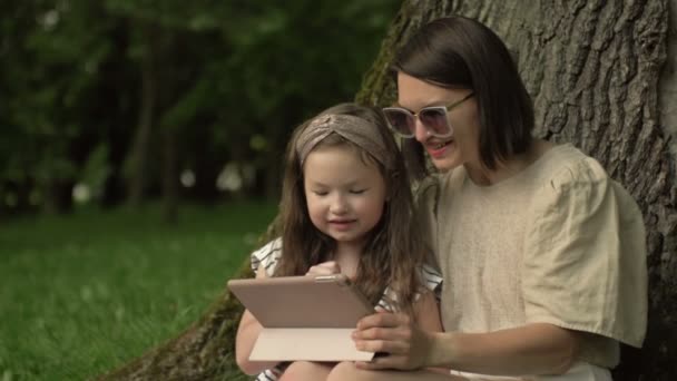 Jovem mulher com uma pequena filha está sentada no parque sob uma grande árvore velha e olhando para a tela do tablet com prazer. — Vídeo de Stock