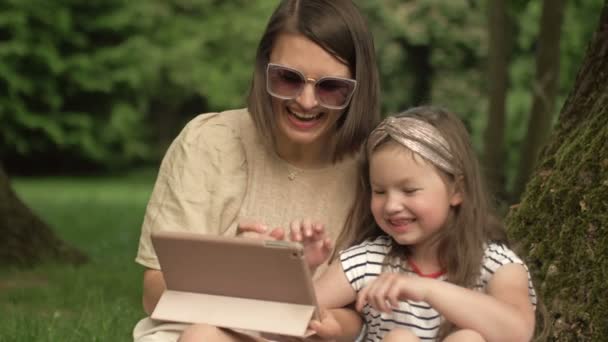 Young woman with a little daughter is sitting in the park under a large old tree and looking at the tablet screen with pleasure. — Stock Video