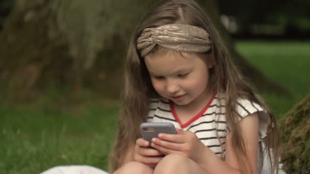 Close-up. Menina bonito 6-7 anos se senta na grama em um parque de verão com um telefone celular em suas mãos. A criança está focada em um jogo de computador. Passatempos prediletos de crianças modernas. — Vídeo de Stock