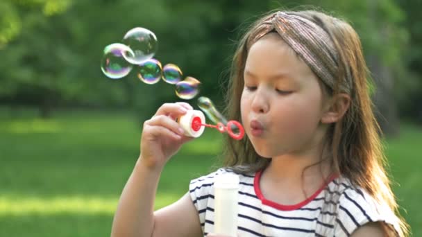 Забавная беззубая дошкольница с энтузиазмом пускает мыльные пузыри. На фоне свежей летней зелени. — стоковое видео