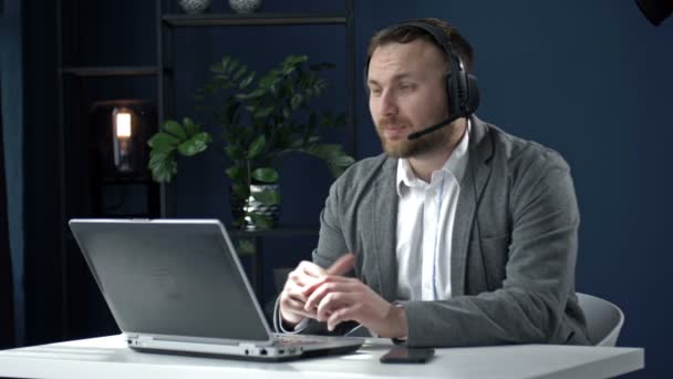 Молодий бізнесмен в гарнітурі сидить за офісним столом перед ноутбуком. Чоловік розмовляє з партнером через відеодзвінок . — стокове відео