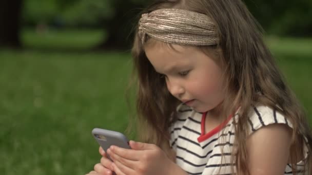 Yakın plan. 6-7 yaşlarında şirin bir kız, elinde cep telefonuyla bir yaz parkında çimlerin üzerinde oturuyor. Çocuk bir bilgisayar oyununa odaklanmış. Modern çocukların hobileri. — Stok video
