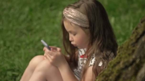 Detailní záběr. Roztomilá dívka 6-7 let sedí na trávě v letním parku s mobilním telefonem v ruce. Dítě se zaměřuje na počítačovou hru. Koníčky moderních dětí. Boční pohled. — Stock video