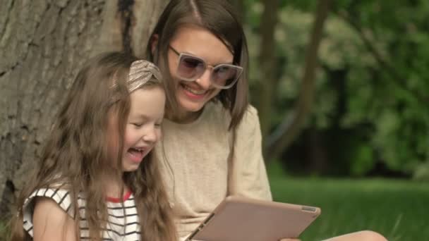 Mujer joven con una hija pequeña está sentado en el parque bajo un gran árbol viejo y mirando la pantalla de la tableta con placer. — Vídeos de Stock