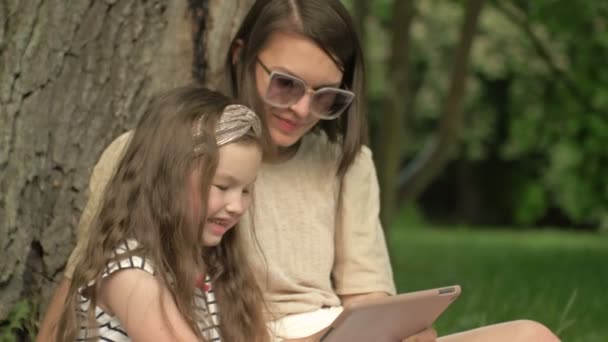 Junge Frau mit kleiner Tochter sitzt im Park unter einem großen alten Baum und schaut genüsslich auf den Tablet-Bildschirm. — Stockvideo