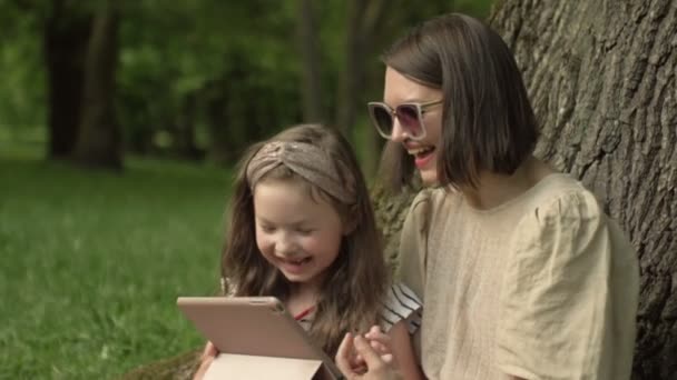 Young woman with a little daughter is sitting in the park under a large old tree and looking at the tablet screen with pleasure. — Stock Video