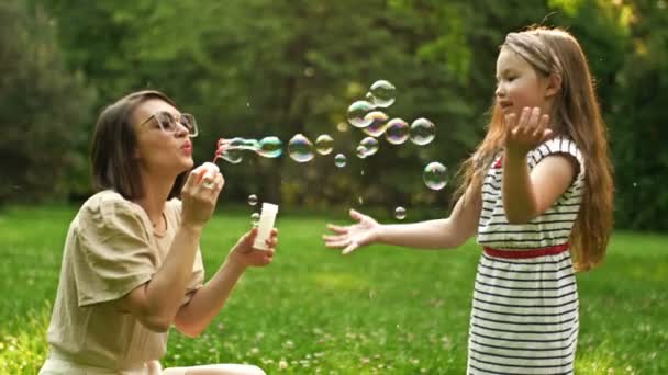 Mooie jonge vrouw zit op groen gras in het park en blaast zeepbellen. Haar 6-jarige dochter vangt graag regenboogbubbels. Gezinsvakantie. — Stockvideo