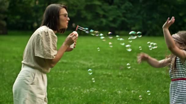 Beautiful young woman sits on green grass in the park and blows soap bubbles. Her 6-year-old daughter happily catches rainbow bubbles. Family holiday. — Stock Video