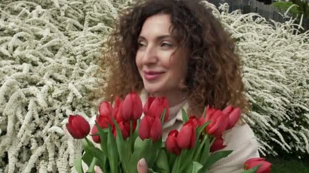 Retrato de uma mulher feliz com um buquê de tulipas vermelhas. Contra o fundo de um arbusto branco florido. Close-up. — Vídeo de Stock