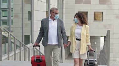 Bavulları olan olgun evli bir çift el ele yürüyor. Yolcuların yüzlerinde tıbbi maskeler var. COVID-19 salgını sırasında seyahat, turizm.