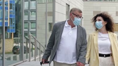 Bavulları olan olgun evli bir çift el ele yürüyor. Yolcuların yüzlerinde tıbbi maskeler var. COVID-19 salgını sırasında seyahat, turizm.