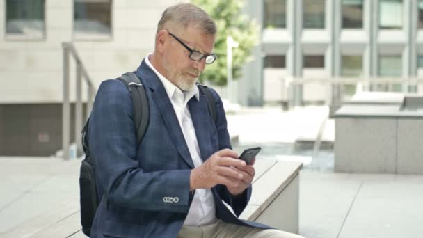 Empresário é um homem de cabelos grisalhos em um terno de negócios sentado perto do escritório se comunica por telefone com colegas via SMS. — Vídeo de Stock