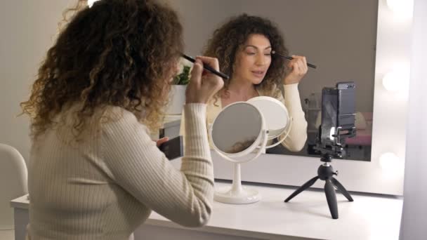 Un bloguero de belleza bien cuidado y de mediana edad habla sobre maquillaje de ojos. Una mujer sentada frente a un espejo y una cámara de video. — Vídeo de stock