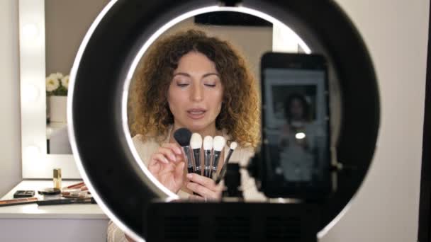 Schöne Beauty-Bloggerin mittleren Alters spricht über die Arten von Make-up-Pinseln und ihren Zweck. Frau sitzt vor einem Spiegel, in dem sich eine Videokamera spiegelt. — Stockvideo