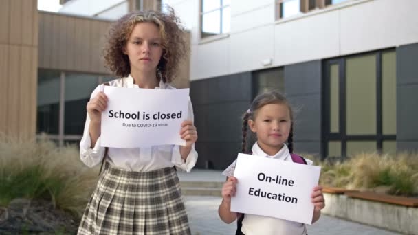 Två skolflickor med plakat står framför skolbyggnaden. En affisch i händerna på en liten flicka ONLINE UTBILDNING. En tonåring flicka innehar en affisch SCHOOL LOSED på grund av Covid-19. — Stockvideo